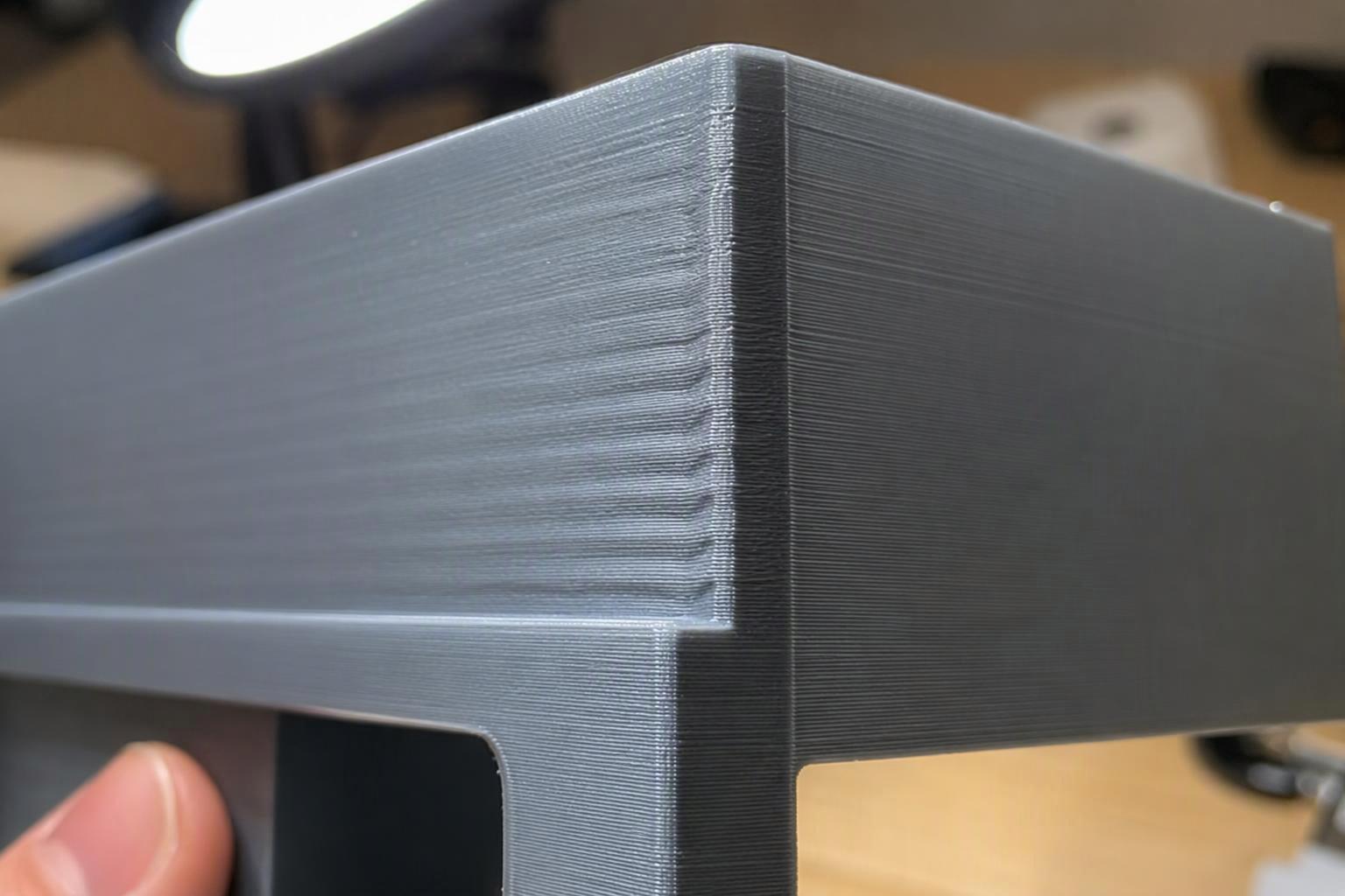 a 3D printed part tilted under a desk lamp showing faint repeating ripples beside a sharp corner