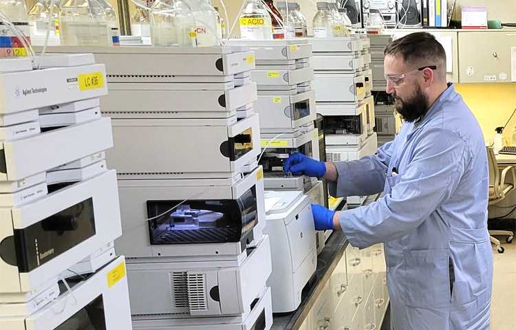 a laboratory bench with an HPLC vial tray and a plate reader in the foreground, sample tubes labeled by timepoint