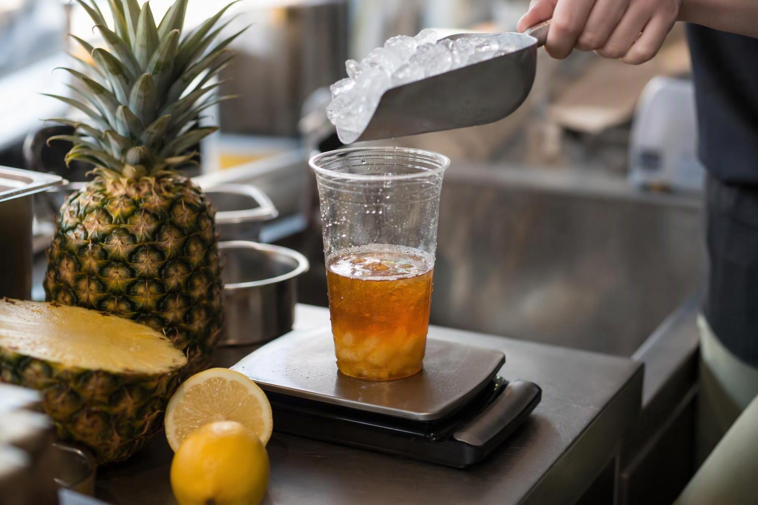 a 16 oz cup on a digital scale mid-build, with the tea base, pineapple puree, and fructose already in and the ice scoop held